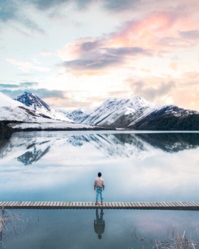 back view of a person standing on wooden planks across the snow capped mountains